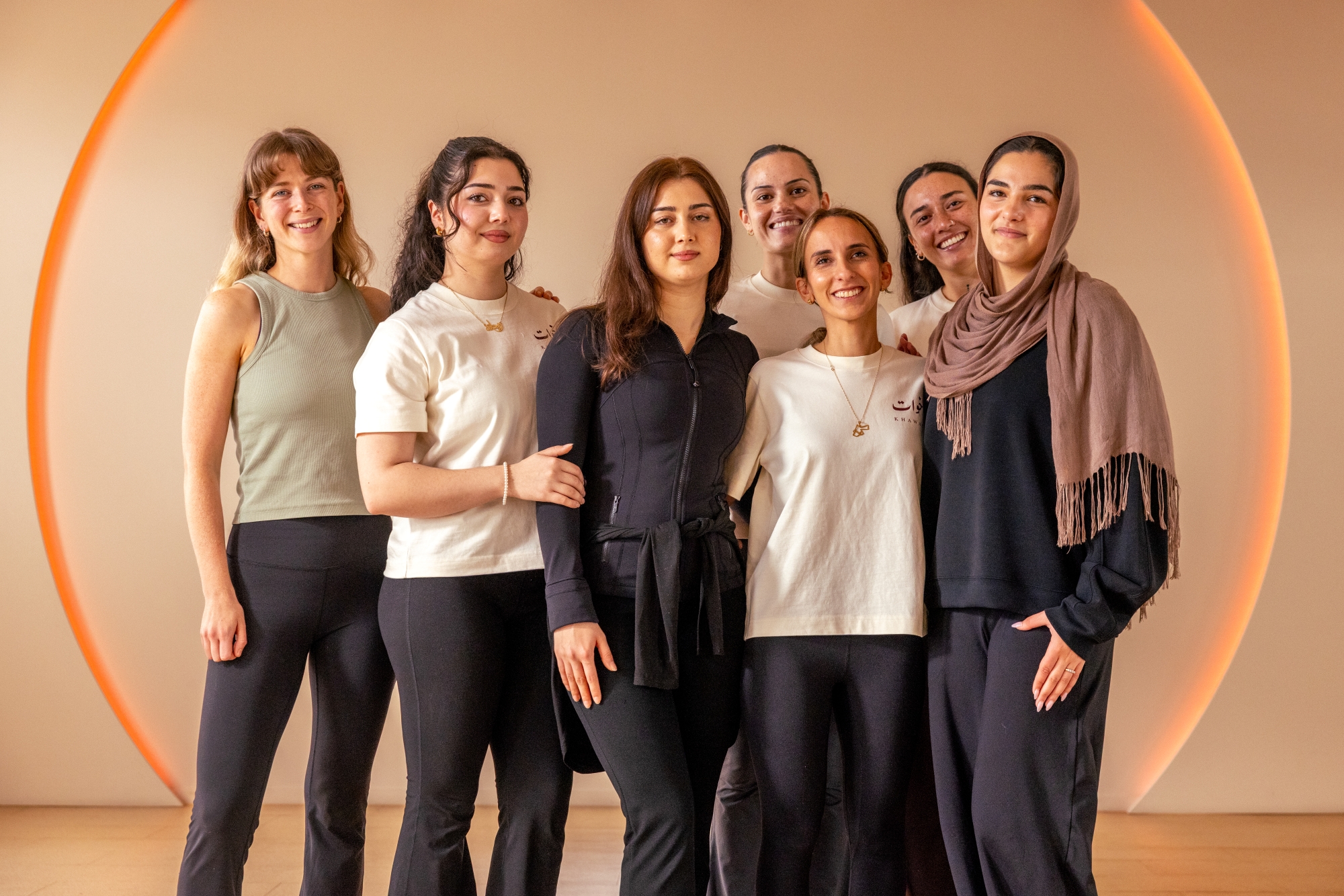 A group of people posing for a photo, they are dressed in a variety of comfortable outfits, stood in a calm space like a yoga studio.