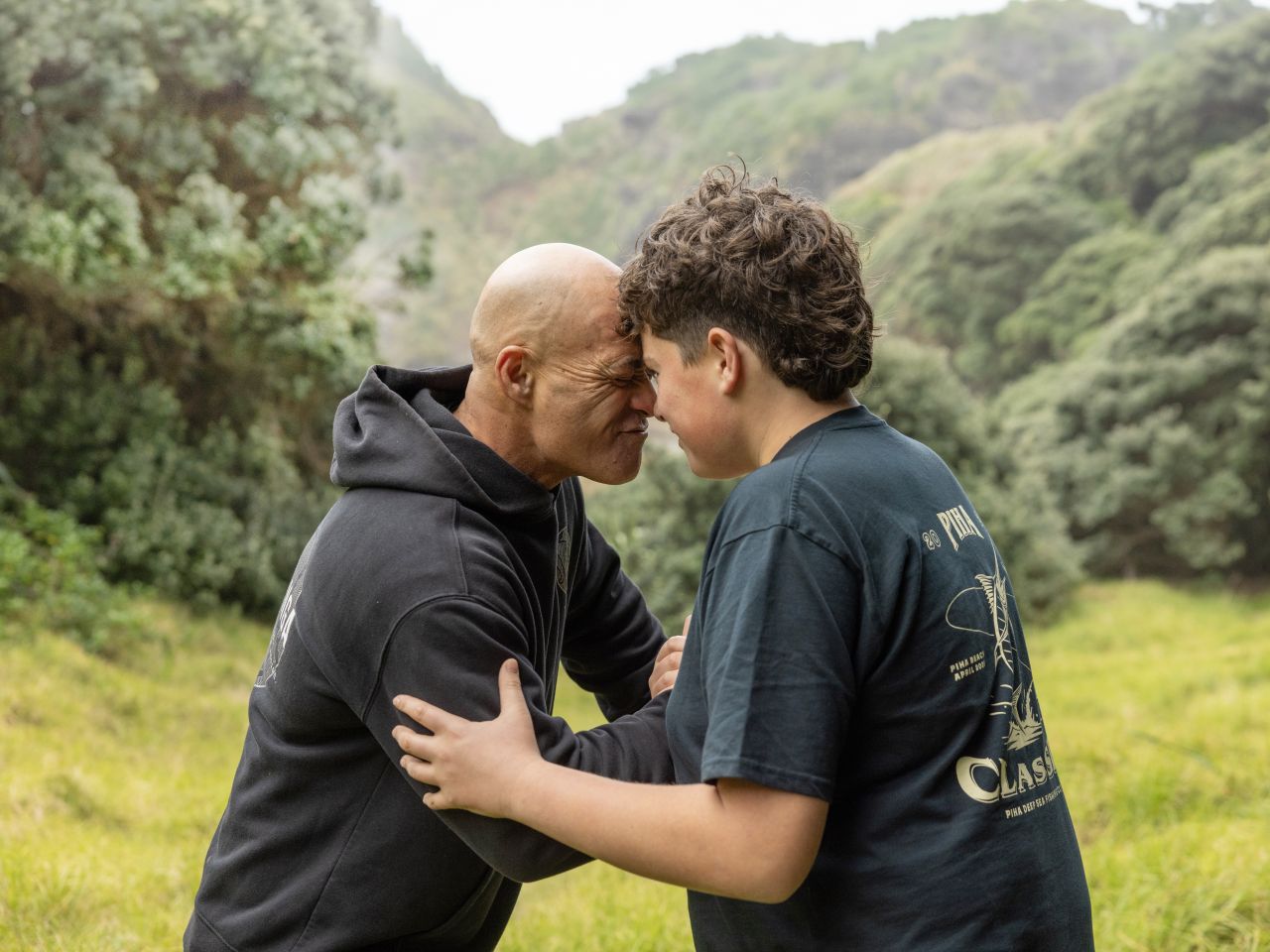 Two people greet each other with hongi, they are amongst a lush green landscape.