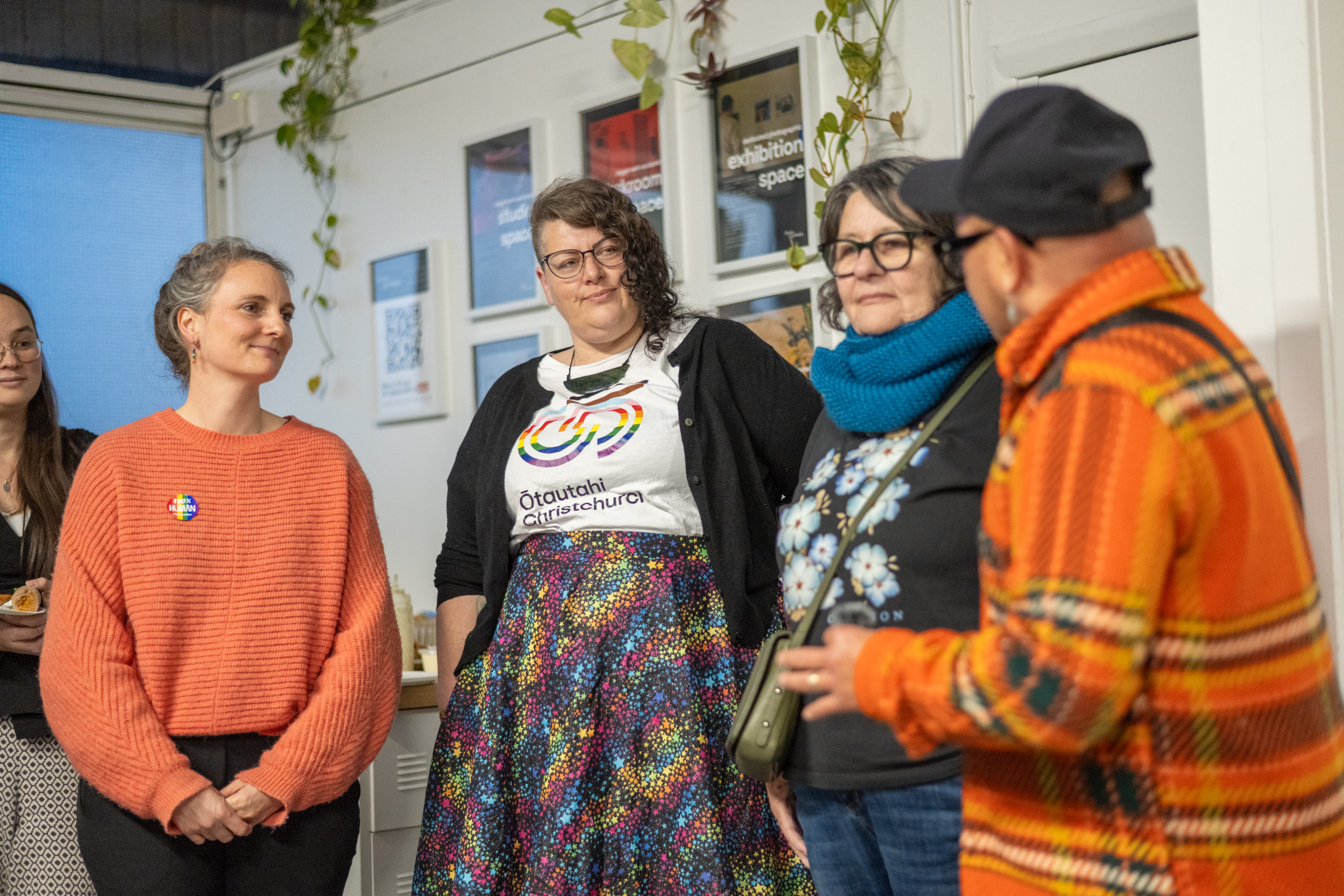 A group of people in an indoor community space, standing and listening to a discussion, there are posters and hanging plants in the background.